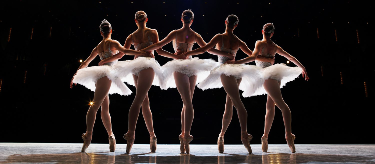 Cinco bailarinas en el escenario, tomadas del brazo, moviéndose como una