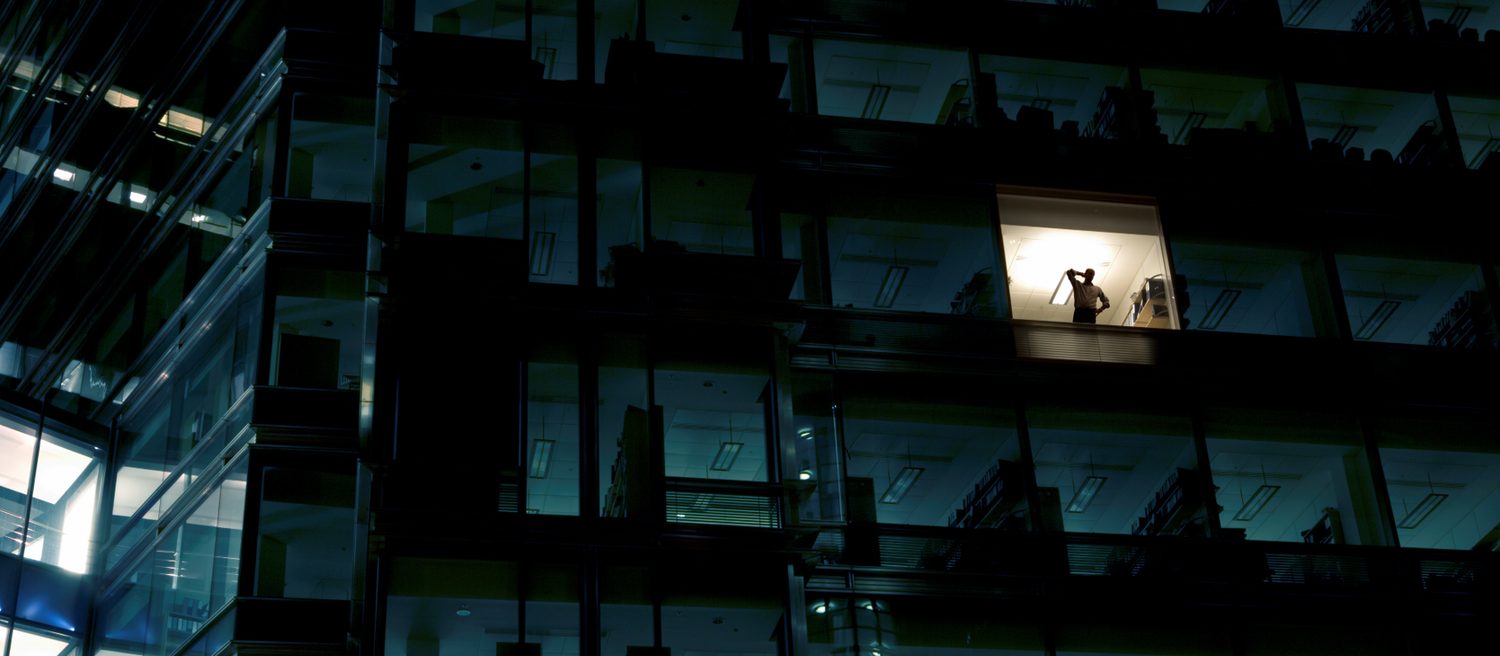 Un edificio de oficinas de noche, con una sola ventana encendida y una persona dentro